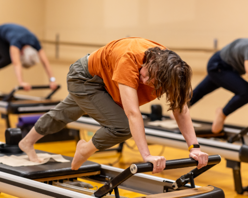 An older woman in an orange shirt uses a reformer pilates machine in Mizzourec doing an advanced pose
